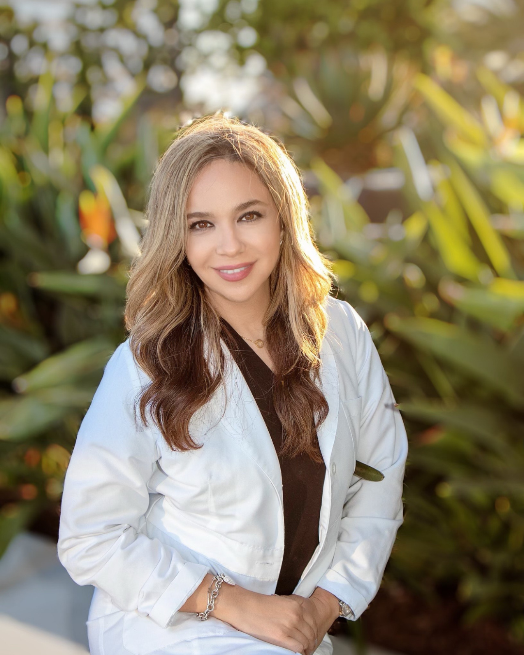 Professional aesthetic injector in a white coat, smiling warmly in a lush outdoor setting, embodying confidence and expertise in aesthetic treatments.